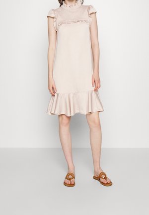 Nainen pukeutuneena vaaleanpunaiseen hihattomaan mekkoon, jossa on röyhelökaulus ja -helma, yhdistettynä beigeihin avosandaaleihin.