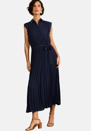 Vestido midi sin mangas de color azul marino con pliegues, botones en la parte delantera, bordes en forma de ola, un cinturón de lazo y un diseño fluido. Llevado con tacones de tiras.