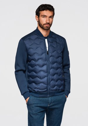 Homme aux cheveux courts et foncés avec une barbe, portant une veste bomber matelassée bleu marine et un jean bleu, debout les mains dans les poches.