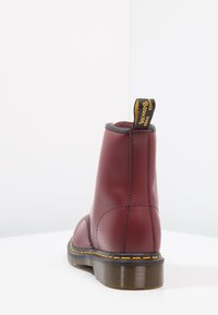 Bottine en cuir bordeaux avec coutures jaunes, semelle noire et languette en tissu avec texte jaune à l'arrière, vue de dos.