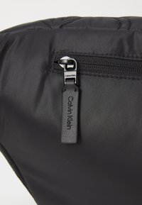 Zwarte gevoerde tas met een gestikte textuur, voorzien van een metalen rits en een leren label met de opdruk "Calvin Klein."