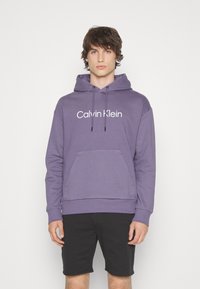 Felpa in cotone viola con tasca a marsupio frontale e cappuccio con coulisse. Caratterizzata dal logo bianco "Calvin Klein" sul petto. Pantaloni corti neri.