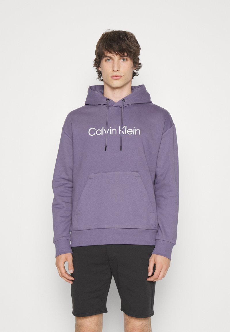 Felpa in cotone viola con tasca a marsupio frontale e cappuccio con coulisse. Caratterizzata dal logo bianco "Calvin Klein" sul petto. Pantaloni corti neri.