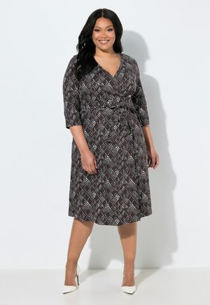 Donna con lunghi capelli neri indossa un vestito nero al ginocchio con pois geometrici bianchi e tacchi alti bianchi, in piedi su uno sfondo bianco.