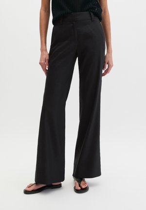 Schwarze, hoch taillierte, weit geschnittene Hose, getragen mit schwarzem ärmellosem Oberteil und schwarzen Flip-Flop-Sandalen auf weißem Hintergrund.