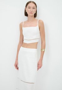 Weißes Leinen-Crop-Top mit dünnen Trägern, versehen mit einem strukturierten Band, und einem passenden weißen Rock mit Spitze am Saum, getragen mit goldenen Armbändern.