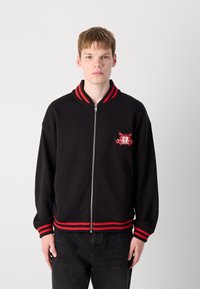 Black zip-up jacket with red and black ribbed collar and cuffs, featuring an embroidered design on the left chest area. Cotton blend material.
