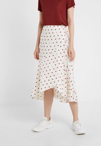 Gonna avvolgente in tessuto bianco con pois rossi, con un orlo alto-basso e chiusura laterale con fiocco, abbinata a una maglietta bordeaux e sneakers bianche.