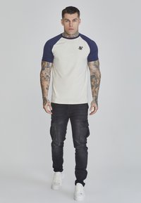 Kurze Ärmel in Creme und Marineblau Raglan-T-Shirt mit kleinem Logo auf der Brust, kombiniert mit schwarzen Cargo-Hosen und weißen Sneakers.
