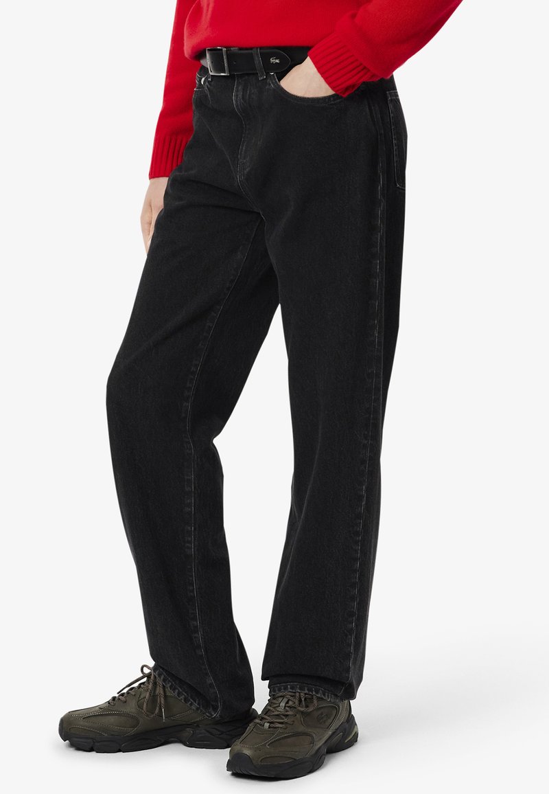 Pantalones de mezclilla negra con un corte relajado, que presentan costuras visibles, bolsillos y un cinturón trenzado. Combinados con calzado oscuro y texturizado.