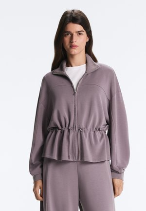 Nő mauve színű, cipzáras zakóban, ami összegyűrt derékrésszel és hozzáillő, bő szárú nadrágban, egyszerű világos háttér előtt áll.