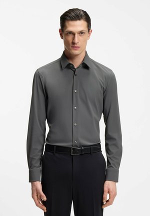 Uomo che indossa una camicia attillata a maniche lunghe grigio scuro con bottoni, infilata in pantaloni neri con cintura nera, in piedi contro uno sfondo bianco.