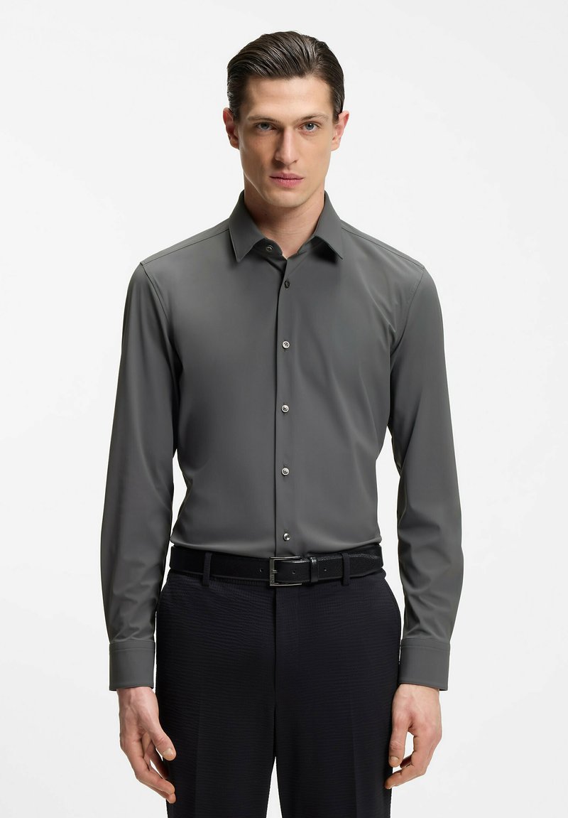 Uomo che indossa una camicia attillata a maniche lunghe grigio scuro con bottoni, infilata in pantaloni neri con cintura nera, in piedi contro uno sfondo bianco.