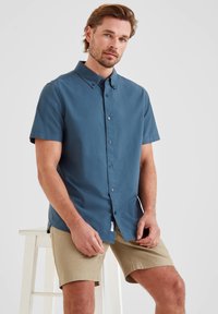 Short-sleeve button-up shirt in blue fabric, featuring a collar, front buttons, and a small logo tag at the hem. Worn with beige shorts.