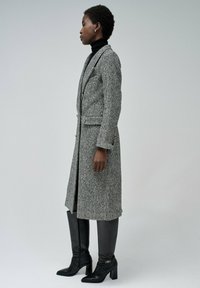 Cappotto in lana a motivo pied-de-poule grigio e nero, con vestibilità sartoriale, due tasche frontali e bottoni dorati. Abbinato a stivali neri alti fino al ginocchio.