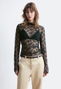 Schwarzes, durchscheinendes Oberteil mit langen Ärmeln und floralen Spitze-Muster über einem schwarzen Bralette, kombiniert mit hellbeigen Hosen und einem dunklen Gürtel.