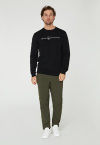 Schwarzer Sweatshirt mit "Sail Racing"-Logo, Rundhalsausschnitt, langen Ärmeln, kombiniert mit olivgrünen Hosen und weißen Sneakers. Minimalistisches Design.
