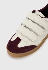 Sneaker with a cream leather upper, burgundy suede toe and heel, textured Velcro straps, and a gum rubber sole.