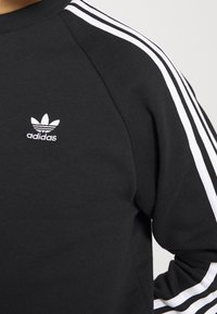 Svart sweatshirt med vita tre-rands accenter på axlarna. Har en broderad vit adidas-logotyp på bröstet. Tillverkad av slät tyg.