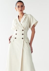 Beige mähkiv kleit tekstureeritud kangast, millel on topelt rinnatükk suurte nööpidega, lühikesed varrukad ja küljetaskud.