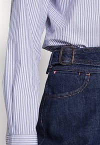 Randig blå och vit långärmad skjorta i kombination med mörkblå jeans. Har en silverfärgad spänne och kontrasterande sömmar.