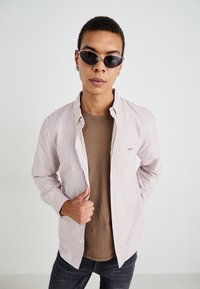 Striped button-up shirt in white and red, worn over a tan t-shirt, paired with black sunglasses. Dark denim jeans complete the look.