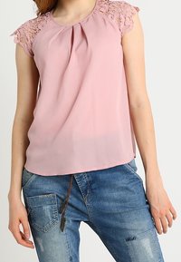 Blusa rosa con maniche in pizzo, davanti plissettato e texture morbida. Abbinata a jeans in denim blu scoloriti.