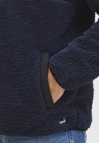 Navy blue fleece pullover with a textured surface, featuring black side pockets and a small logo detail near the hem.