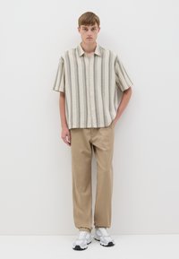 Camicia a maniche corte con bottoni, a strisce verticali beige e grigie, abbinata a pantaloni marroni chiari e sneakers grigie. Sfondo semplice.