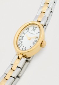 Timex ur med et oval guld-tonet kabinet, hvid urskive, sorte romerske tal, guld h�ænder og et bicolore metalrem i sølv og guld.