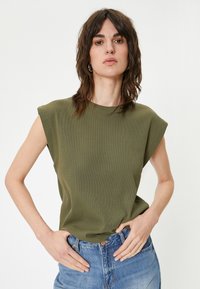 Olivengrøn, ærmeløs top med høj hals og en struktureret overflade. Iført med højtaljede blå jeans, hænderne på hofterne.
