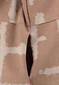 Tela color tan con una textura suave y lisa, salpicada de manchas blancas irregulares. El diseño incluye una abertura central y un escote redondeado.