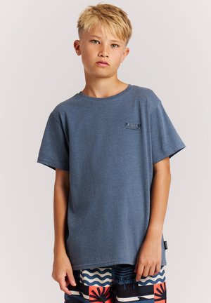 Blond jongetje met een neutrale uitdrukking, gekleed in een blauw T-shirt en kleurrijke, patroonrijke korte broek, staand tegen een effen lichte achtergrond.