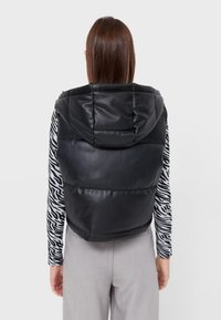 Schwarze Pufferweste mit Kapuze, aus glattem, kunstlederähnlichem Material. Getragen über einem gestreiften Langarmoberteil und kombiniert mit hellen Hosen.