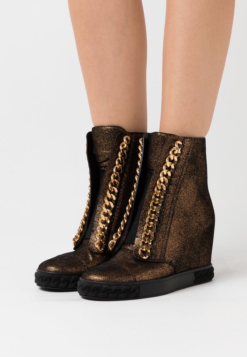 Chevilles portant des baskets compensées noires avec une texture scintillante et des ornements en chaîne dorée proéminents le long de la languette.