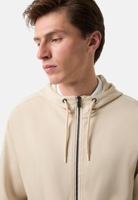Krémová mikina na zip s nastavitelnými šňůrkami, hladkým povrchem, minimalistickým designem a kontrastním zipem. Model ji nosí s bílým tričkem.