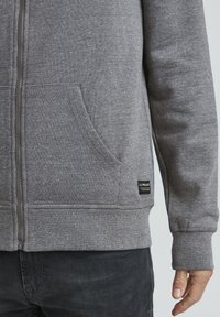 Siva zip-up dukserica od mekog flisa s prednjim džepom i detaljima na crnom etiketi. Sadrži rebraste manžete i donji rub za čvrsto prianjanje.