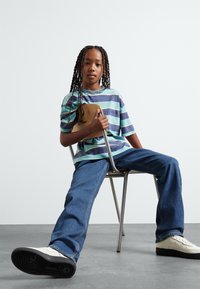Striped t-shirt in teal and navy, blue jeans, and white sneakers. The child holds a skateboard while seated on a metal chair.