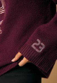 Burgundin neulepaita, jossa on joustinneulehihansuut, brodeerattu logo ja valkoinen numero-osio hihassa.