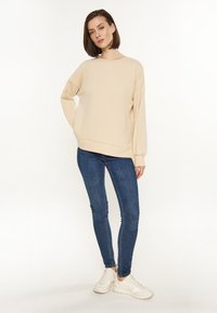 Beige garbós pulóver, hosszú ujjakkal és laza szabással, kék skinny farmernadrággal és fehér sportcipővel párosítva, világos háttér előtt.