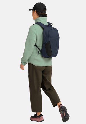 Persona con cappellino nero, giacca in pile verde, pantaloni marroni, scarpe da trekking rosa e nere e zaino blu navy che cammina di spalle su sfondo bianco.