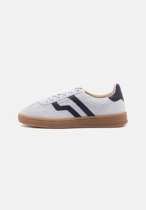 Low-top suede sneaker in light gray with dark blue stripes, gum rubber sole, and beige interior lining, side view on white background.