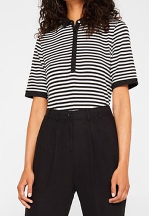 Frau trägt ein schwarz-weiß horizontal gestreiftes kurzärmeliges Shirt und hoch taillierte schwarze Hose, steht mit entspannten Armen seitlich.