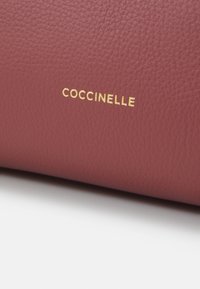 Burgundi bőr kézitáska, texturált felülettel és a „COCCINELLE” feliratú, arany dombornyomott logóval kiemelten elhelyezve.