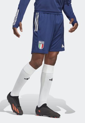 Homme portant un short de football italien bleu, des chaussettes blanches montantes jusqu'aux genoux et des crampons noirs avec des rayures orange, debout, les jambes légèrement croisées.
