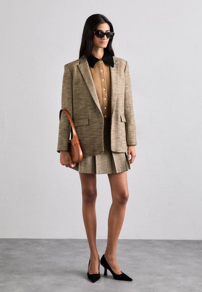 Moteris vilkinti margintą švarką ir sijoną, šviesiai rudą marškinėlį su juodu apykakle, juodus aukštakulnius, saulės akinius ir laikanti rudą rankinę.