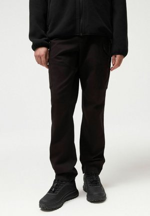 Osoba ubrana w czarne spodnie bojówki, czarną polarową kurtkę i czarne buty do chodzenia po górach, stojąca na jasnym, jednolitym tle.