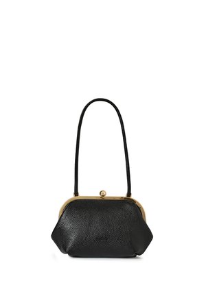 Vrouw met krullend donker haar, gekleed in een lange crème coltrui-jurk, met een zwarte handtas, staand voor witte deuren.