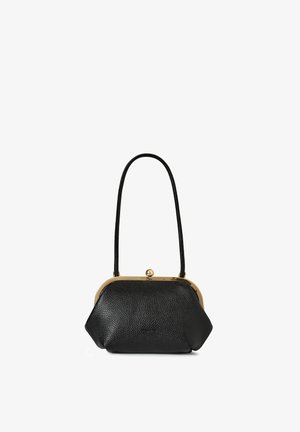 Kobieta o kręconych ciemnych włosach, ubrana w długą kremową sukienkę z golfem, trzymająca czarną torebkę, stojąca przed białymi drzwiami.