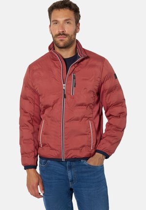 Mann trägt eine rostrot gesteppte Jacke mit Reißverschlusstaschen und blaue Jeans, steht mit einer Hand in der Tasche vor einem schlichten weißen Hintergrund.